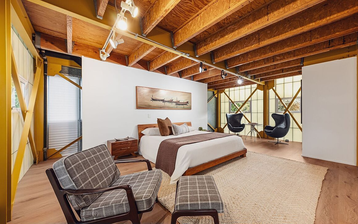 A character loft bedroom with exposed wood-beam ceiling, mid-century plaid chairs, and yellow X-bracing on the walls.
