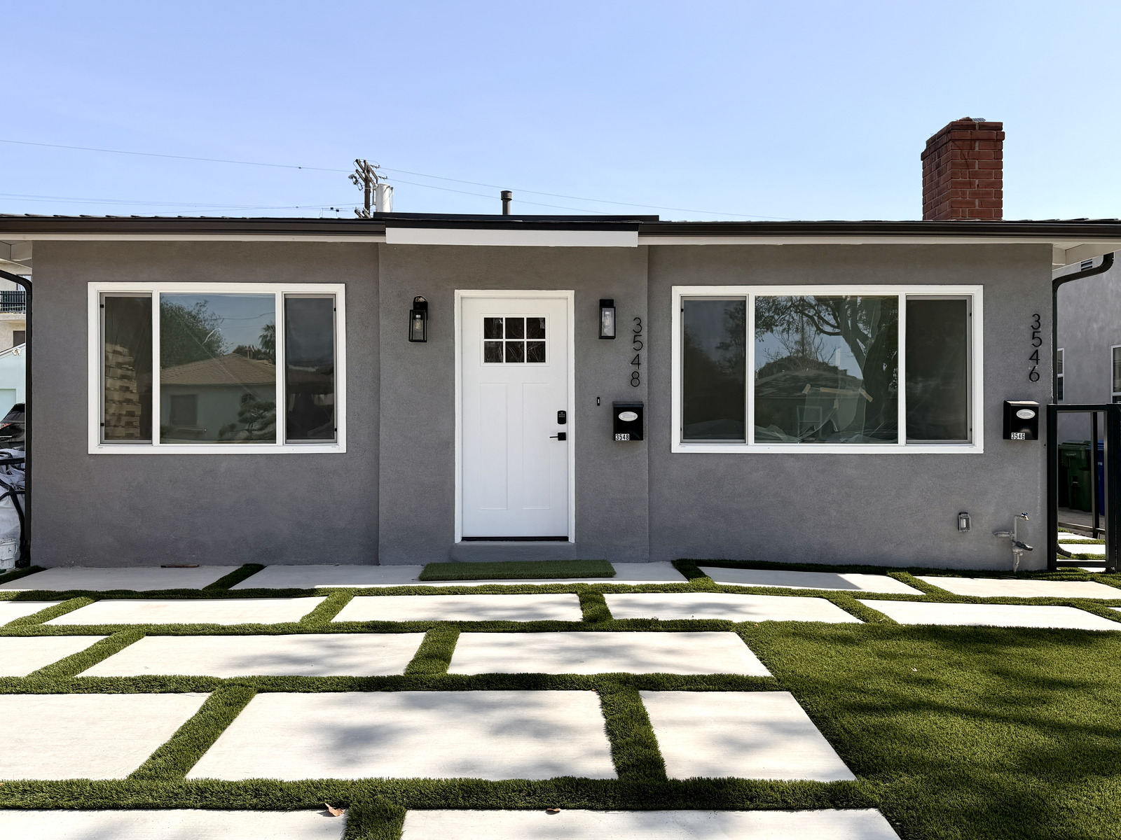 Completed single-story detached ADU with gray stucco façade, white craftsman door, and paver-and-grass hardscape.