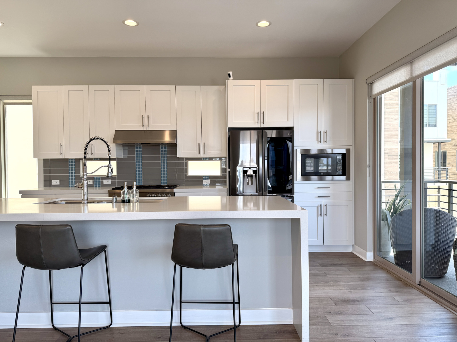 Bright modern kitchen with white shaker cabinets, stainless appliances, quartz waterfall countertop, and leather counter stools.